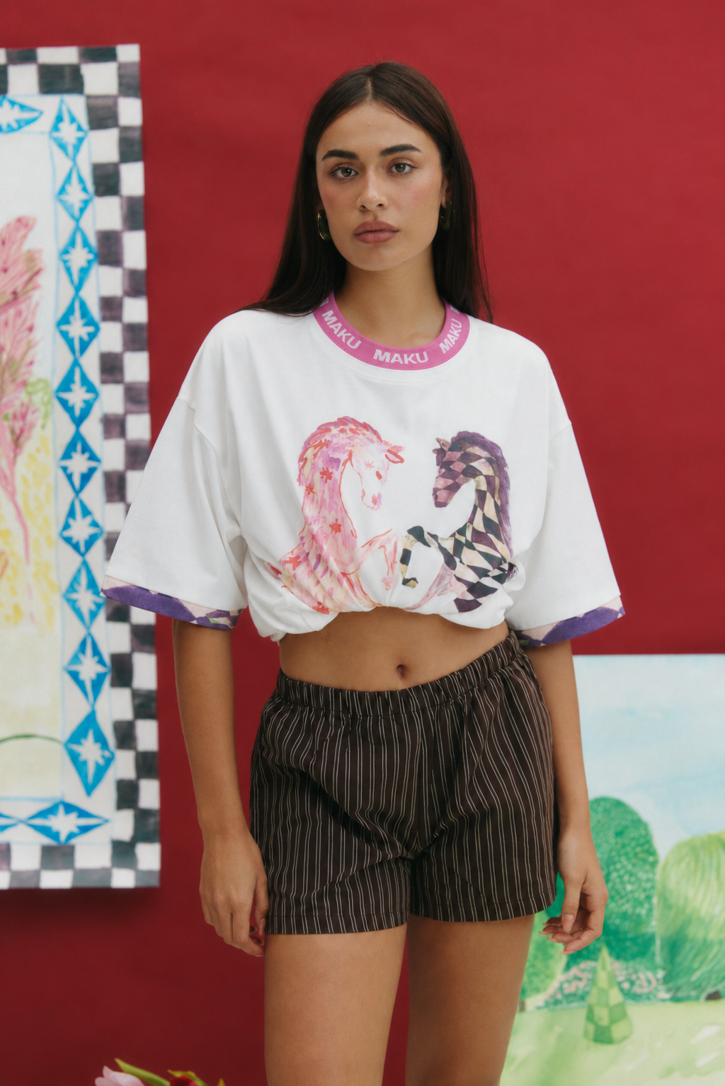 A woman modeling a white cropped graphic t-shirt with pink and purple details, paired with dark pinstriped shorts, set against a red wall with colorful art prints.