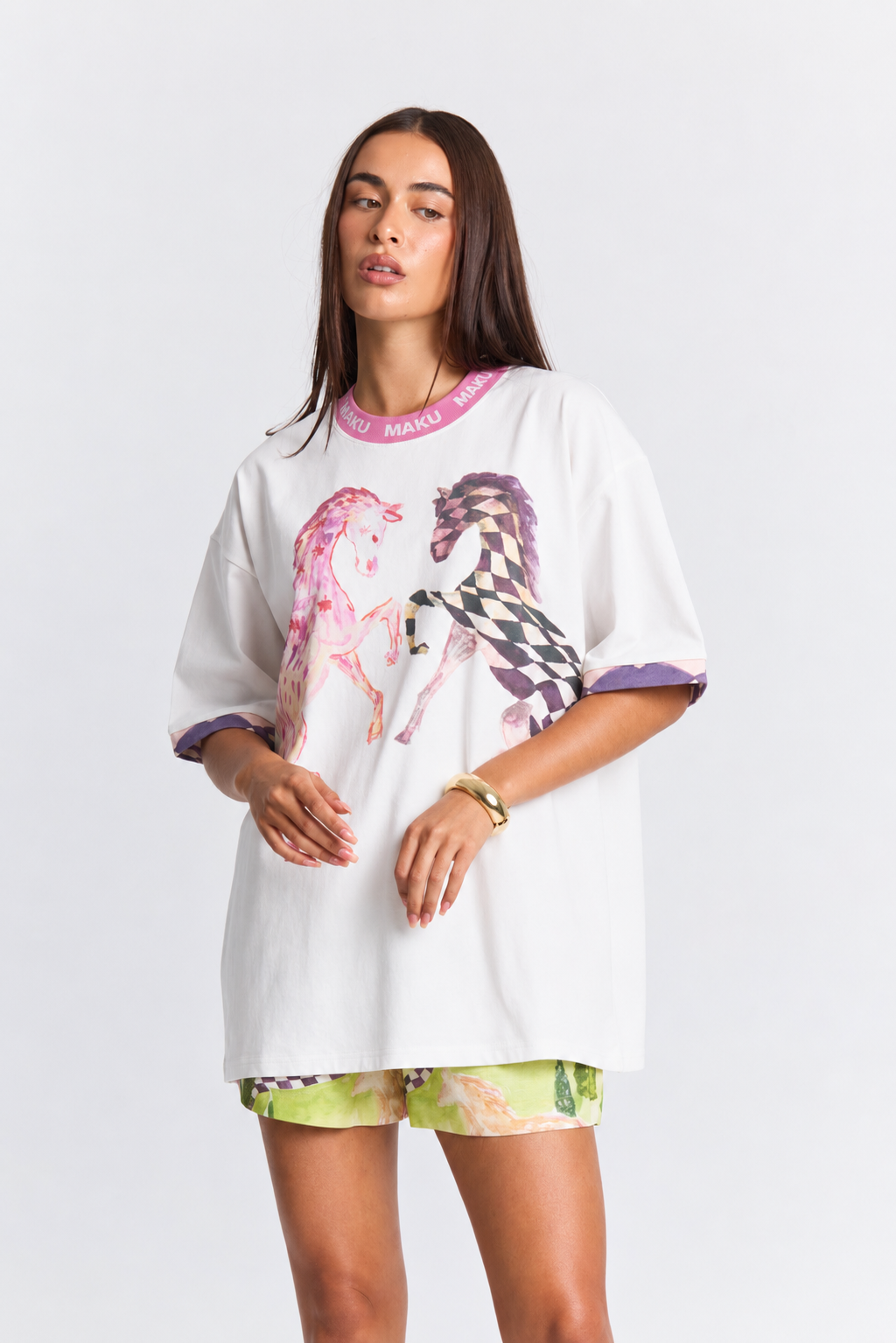 A woman modeling an oversized white T-shirt with a pink and purple horse graphic, pink trim, and green floral shorts against a light gray studio background.